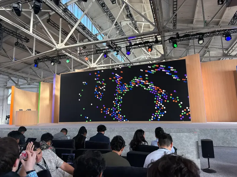 Attendees sit facing a large stage screen displaying a pattern made of colored d