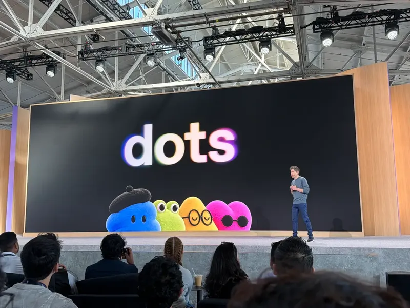 A presenter stands onstage before an audience beside a giant screen displaying “