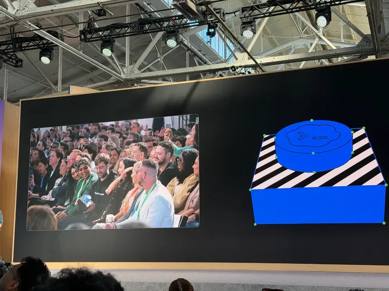 A large presentation screen at a conference shows an audience seated in rows on 