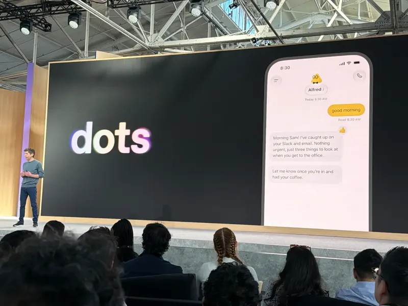 A presenter stands before an audience onstage beside a large screen displaying t